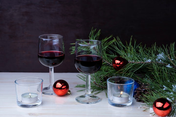 Two glasses with red wine stand on a wooden table. Around them lie pine branches covered with snow, Christmas balls and candles.