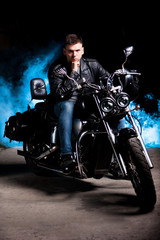 Fototapeta na wymiar Cute biker in leather jacket sits on a motorcycle in blue smoke 