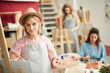 Young creative woman with palette and paintbrush painting in studio of arts