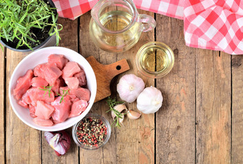 VIew from above of raw pork meal in a bowl, garlic, onion, rosemary in a pot, pepper, jug with oil and checkered red tablecloth in the background