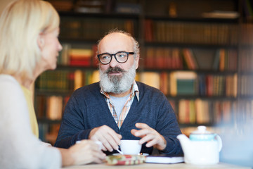 Senior bearded man talking to his wife or colleague by cup of coffee in cafeteria