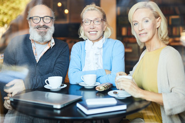 Fototapeta na wymiar Three happy seniors in smart casual looking at you while sitting by table and relaxing after tea-time