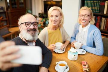 Group of friendly seniors making selfie while sitting by table in cafe during tea time