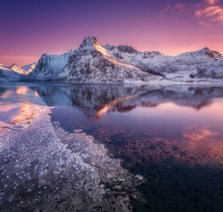 Aerial view of snowy mountains, blue sea with frosty coast, reflection in water and purple sky at colorful sunset in Lofoten islands, Norway. Winter landscape with snow covered rocks, fjord with ice