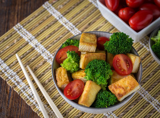 Grilled tofu with broccoli and tomatoes in white bowl on bamboo pad with chopsticks on the left side.