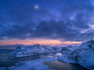 Fototapeta na wymiar Aerial view of snowy mountains, sea, colorful cloudy sky at night in Lofoten islands, Norway. Winter landscape with snow covered rocks and seacoast and sunset sky. Top view of Norwegian Fjords at dusk