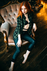 A beautiful slender girl with long red hair in leather pants and an unbuttoned plaid shirt is sitting in a chair