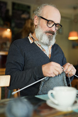 Bearded mature man concentrating on knitting warm winterwear by cup of tea
