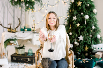 Merry Christmas, New Year, family, holidays concept. Happy young Caucasian woman in jeans and knitted white sweater sits in the armchair in front of the Christmas decorations and holds bengal light