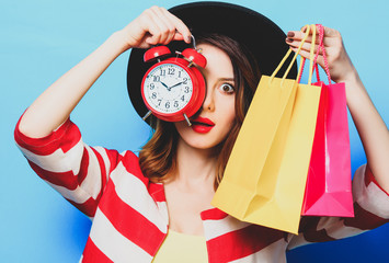 Portrait of young surprised red-haired white european woman in hat and red striped shirt with alarm clock and shopping bags on blue background