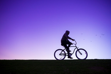 Fototapeta na wymiar Silhouette of man in hoodie riding a beach cruiser bicycle against a glowing purple sky
