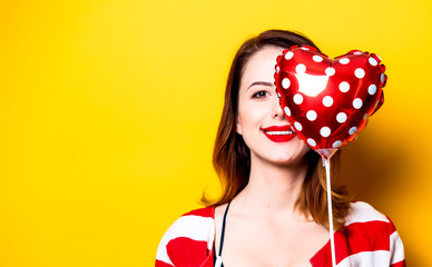 Portrait of beautiful young smiling red-haired white european woman in red striped shirt with heart shape toy on yellow background