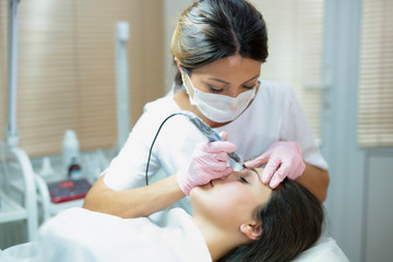 Make-up. Beautician in pink gloves doing eyebrow tattoo on woman's face. Closeup of specialist doing eyebrow tattooing for female.