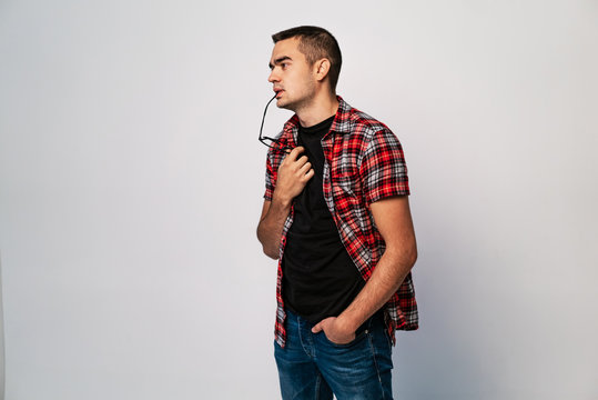 Young male with eyeglasses to lips thinking a deep thought on white background. A portrait of handsome man in shirt and jeans with glasses in his hand and mouth. Concept problem solving. - Powered by Adobe
