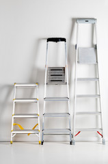 Three aluminum folding metal step ladders leaning against white plaster wall © Shawn Hempel