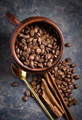 Roasted coffee beans in old clay mug, cinnamon sticks and spoon on gray background. Top view