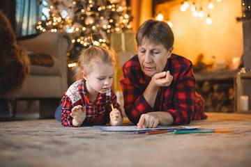 Fototapeta na wymiar Grandma and granddaughter lie on the carpet in front of the Christmas tree and draw. Family christmas concept.