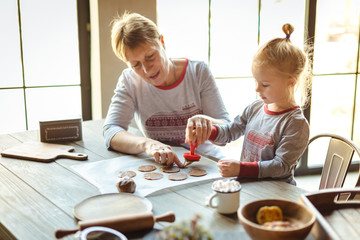 Grandmother and granddaughter in the morning in the same pajamas together bake Christmas cookies-stamps on the test. The family cozy Christmas concept. Selective focus.