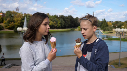 Two teenage kids / young adults enjoying eating strawberry, chocolate and vanilla ice cream cones while laughing, smiling and chatting to each other on a hot sunny day outside by a beautiful lake