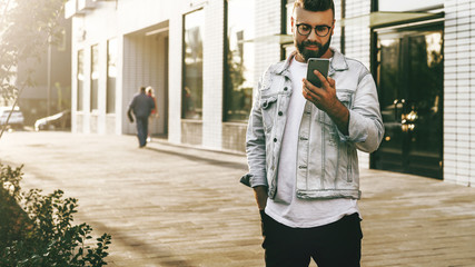 Fototapeta premium Handsome smiling hipster businessman with beard, in denim jacket, trendy glasses walks around city and using smartphone.