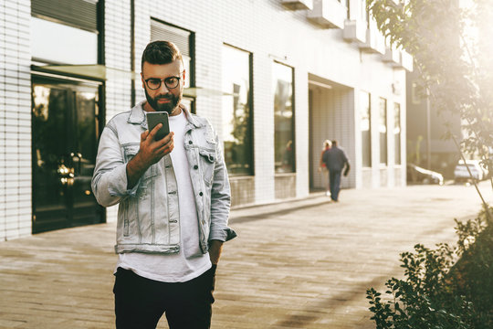 Handsome smiling hipster businessman with beard, in denim jacket, trendy glasses walks around city and using smartphone.