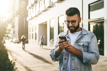 Handsome smiling hipster businessman with beard, in denim jacket, trendy glasses walks around city and using smartphone.