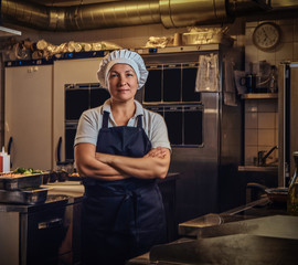 A middle age cook wearing a uniform standing with her arms crossed at restaurant's kitchen. 