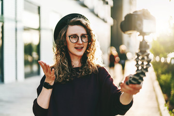 Young female blogger in trendy glasses and hat walks on city street, records herself on video holding camera for tripod.
