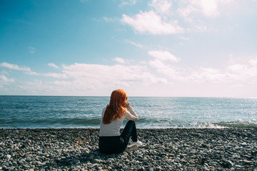 Girl with long ginger hair sitting on the beach and looking into the distance - 235556387