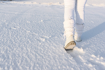 Low angle view of walking away female legs with white snow boots on, in deep snow. Winter background.