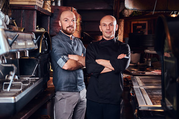 Chef with his assistant posing for a camera with his arms crossed in a restaurant kitchen.
