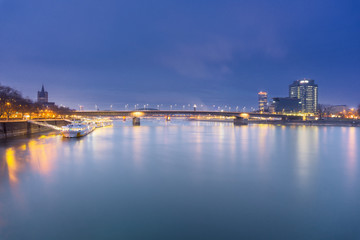 Blick auf den wunderschönen Rhein, Schiffe die sich im Wasser spiegeln, die Deutzer Brücke und die beleuchtete Skyline in Köln bei Nacht,