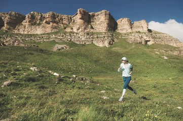 Fototapeta na wymiar Attractive sports girl in a cap and headphones jogging in a picturesque place down the hill near the rocks at sunset. Workout outdoors