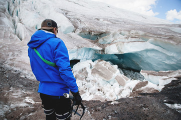 A young guide in a cap with climbing equipment and an ice ax in his hand stands against the background of a crumbling glacier and cracks. The concept of global warming and professional guides