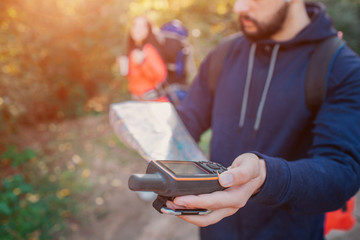 Picture of young bearded man holding satellite phone in hand. There is map in another hand. Young woman stand behind him. She has backpack.
