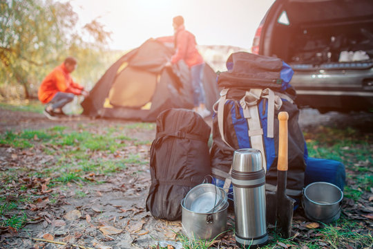 Picture of different expedition equipment. There are backpack with carimate, sleeping bag and dishes. Young man and woman are packing tent. They are at car.