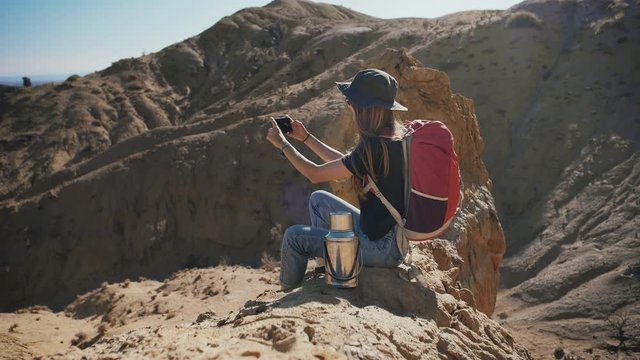 Young traveler hipster female making picture by smartphone sitting in middle of endless desert with mountains rock with a beautiful view of blue sky in vacations or holidays travel to stunning Nature