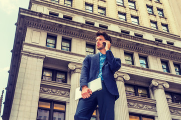 Serious European Businessman traveling, working in New York. Dressing in blue suit, carrying laptop computer, a young guy with beard standing on street with high buildings, talking on mobile phone.