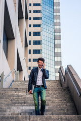 Young professional, arm carrying laptop computer, talking to his family on his mobile phone, walking down stairs outside office building after working, going home. Filtered look with dark blue tint.