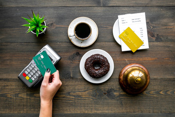 Paypass tech. Contactless payment. Woman's hand brings the bank card to payment terminal near bill, service bell, coffee on dark wooden background top view