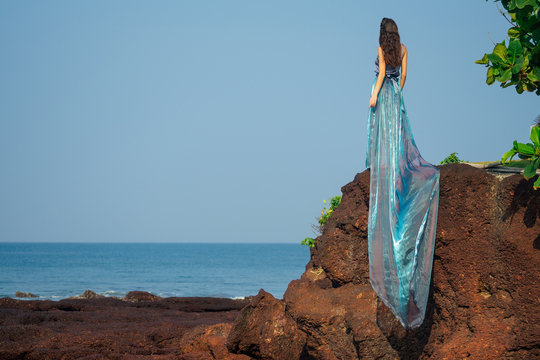 Fashion photo of glamorous brunette woman in a long dress with a train blue chameleon color nymph on the ocean with rocks and waves.