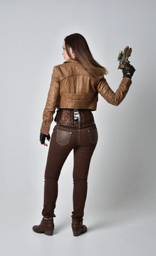 full length portrait of brunette  girl wearing brown leather steampunk outfit. standing pose, holding a gun. on grey studio background.