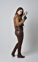 full length portrait of brunette  girl wearing brown leather steampunk outfit. standing pose, holding a gun. on grey studio background.