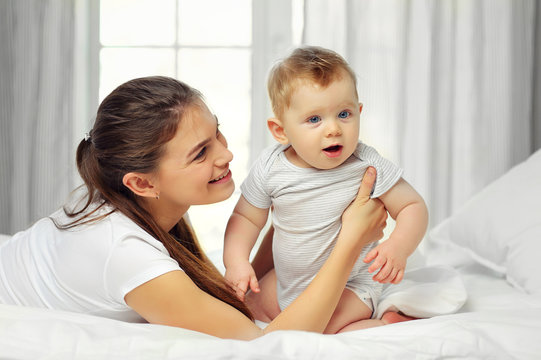 Mom and a charming blue-eyed baby of 10 months in a striped jumpsuit are lying in bed and hugging