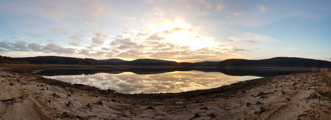 Fototapeta na wymiar scenic panorama view of sunset under a cloudy sky panorama with sun reflection over water - beautiful scene of a lake while golden sunset / sunrise – sun and clouds reflection over panoramic scenery
