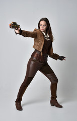 full length portrait of brunette  girl wearing brown leather steampunk outfit. standing pose, holding a gun. on grey studio background. © faestock