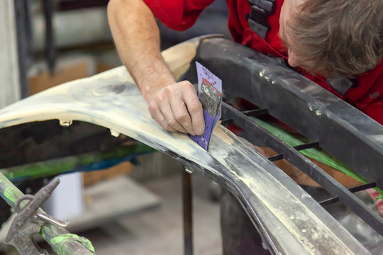 A Man Prepares A Car Body Element For Painting After An Accident With The Help Of Abrasive Paper In A Car Repair Shop. Recovery Bumper After A Collision.