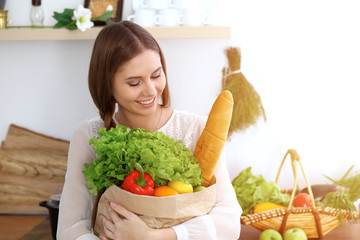 Young happy woman holding paper bag full of vegetables and fruits while smiling. Girl have made shopping and ready for cooking  in kitchen