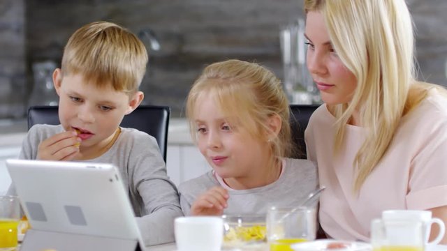 Joyous little girl embracing mom and laughing with elder brother while watching something funny on digital tablet during family breakfast at home