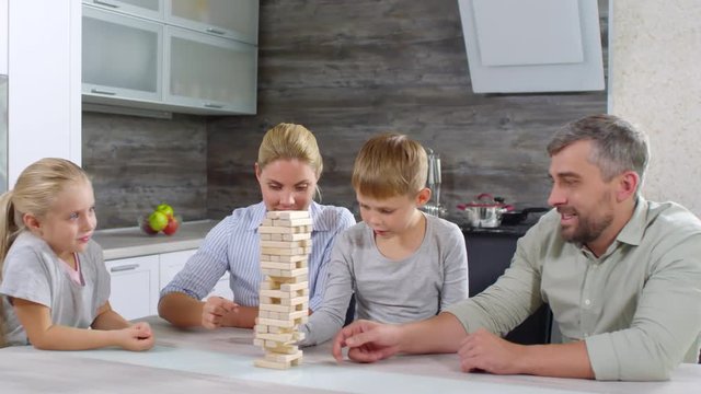 Happy Caucasian family playing table game in the kitchen: mother, father, little daughter and son removing blocks from a tower and laughing when it falls